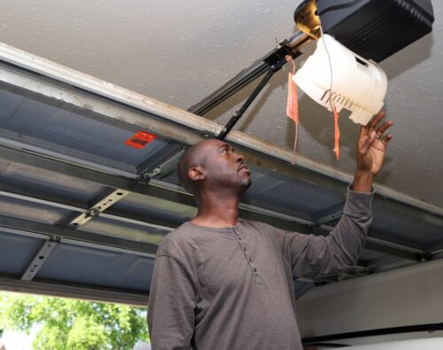 Man reaching up to inspect a ceiling-mounted garage door opener inside a residential garage.