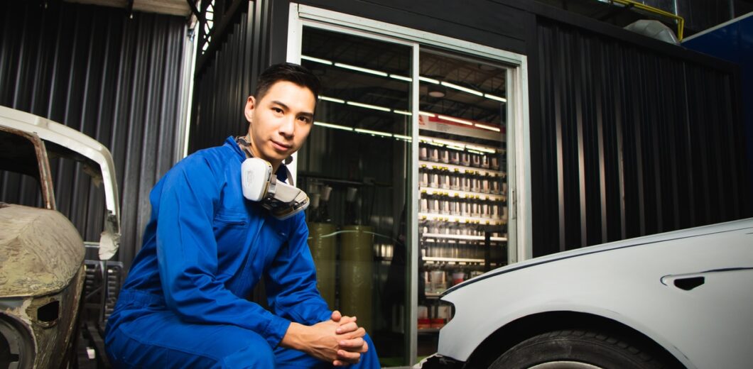Technician in a blue jumpsuit crouching next to a vehicle in an auto repair garage.
