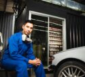 Technician in a blue jumpsuit crouching next to a vehicle in an auto repair garage.