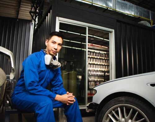 Technician in a blue jumpsuit crouching next to a vehicle in an auto repair garage.