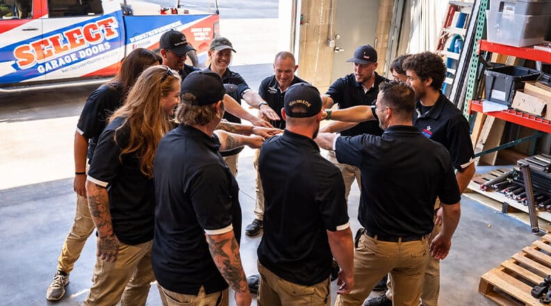 A group of workers in matching uniforms stand in a circle inside a garage, joining hands in the center as part of a team huddle.