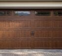 Brown wooden garage door with vertical and horizontal paneling, decorative black hardware, and four small rectangular windows along the top.