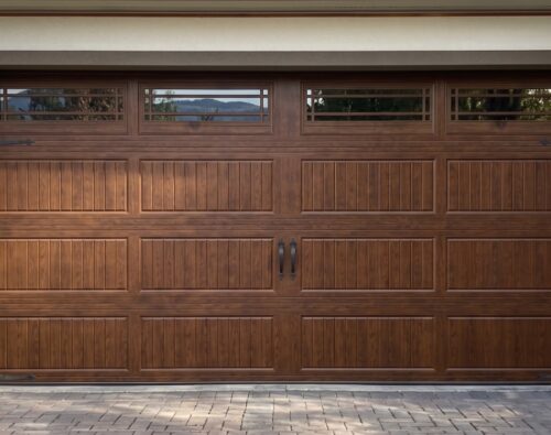 Brown wooden garage door with vertical and horizontal paneling, decorative black hardware, and four small rectangular windows along the top.