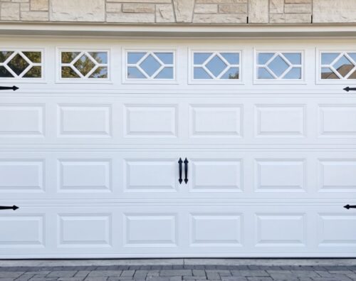 White garage door with four decorative windows at the top, black handles, and black hinge accents, set in a stone exterior wall.
