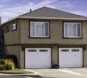 Two-story beige house with two white garage doors, white window shutters, a side staircase, and small landscaped plants in front, on a clear day.