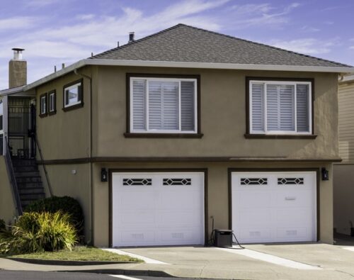 Two-story beige house with two white garage doors, white window shutters, a side staircase, and small landscaped plants in front, on a clear day.