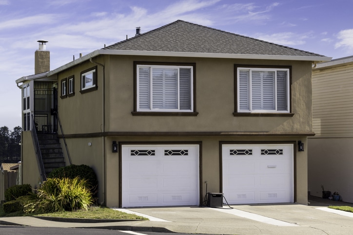 Two-story beige house with two white garage doors, white window shutters, a side staircase, and small landscaped plants in front, on a clear day.