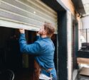 A man in a denim shirt and apron lifts a metal garage-style door to open a storefront or workshop.