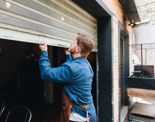 A man in a denim shirt and apron lifts a metal garage-style door to open a storefront or workshop.