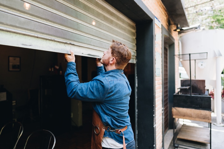 A man in a denim shirt and apron lifts a metal garage-style door to open a storefront or workshop.