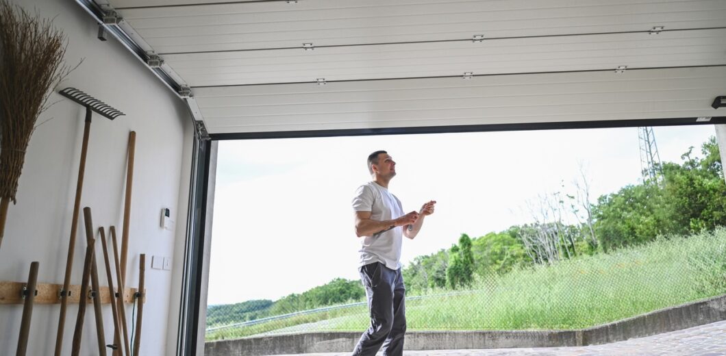 A man in casual clothes walks out of an open garage into a yard with green grass and trees visible in the background.