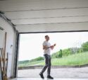 A man in casual clothes walks out of an open garage into a yard with green grass and trees visible in the background.