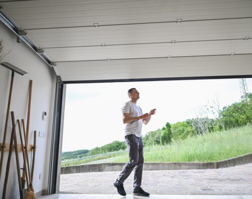 A man in casual clothes walks out of an open garage into a yard with green grass and trees visible in the background.
