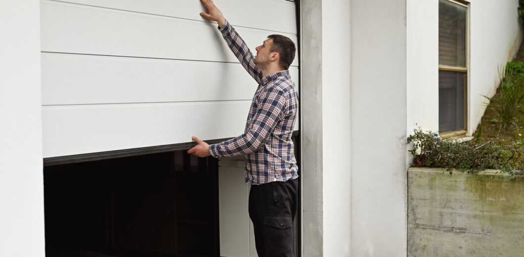 A man in a plaid shirt manually opens or closes a white garage door attached to a white building.