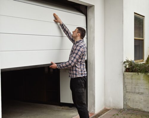 A man in a plaid shirt manually opens or closes a white garage door attached to a white building.