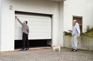 A man opens a garage door while another man stands nearby holding a remote, with a small dog standing on the driveway.