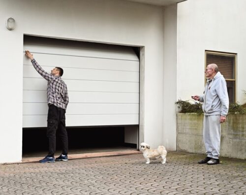 A man opens a garage door while another man stands nearby holding a remote, with a small dog standing on the driveway.