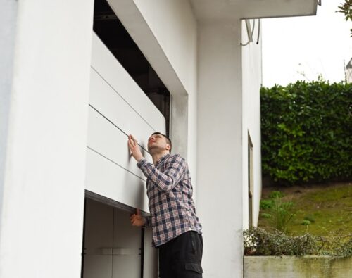 A man in a plaid shirt is manually opening a white garage door attached to a modern house.