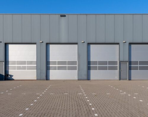 A row of closed industrial garage doors on a gray warehouse building, with an empty paved lot in front and clear blue sky above.