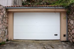 A closed white garage door set between stone walls with a tiled roof above and a concrete driveway in front.