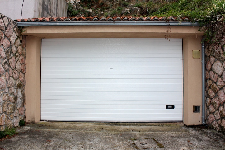 A closed white garage door set between stone walls with a tiled roof above and a concrete driveway in front.