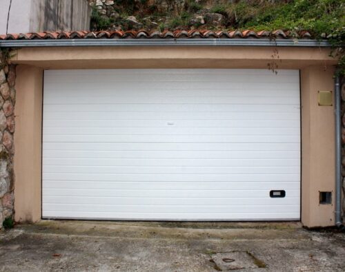 A closed white garage door set in a stone and stucco wall with a tiled roof, on a concrete driveway.