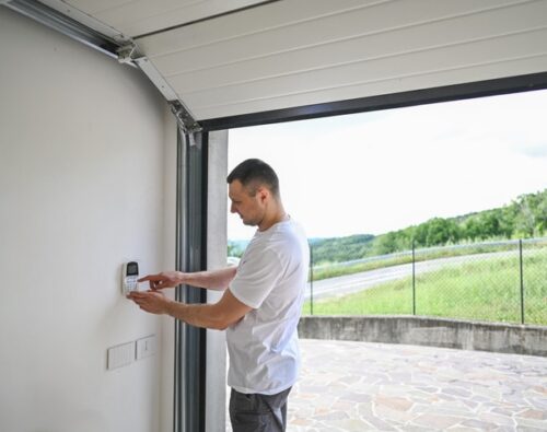 A man stands inside a garage, pressing buttons on a wall-mounted keypad, with the garage door open to an outdoor view.