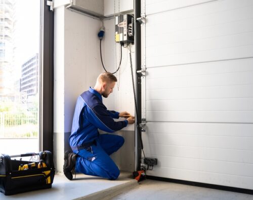 Technician in blue overalls kneels to repair or install the control panel of an automatic garage door, with a tool bag placed nearby.
