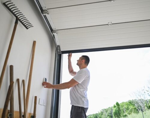 A person in a white t-shirt opens a garage door while standing next to a wall with gardening tools.