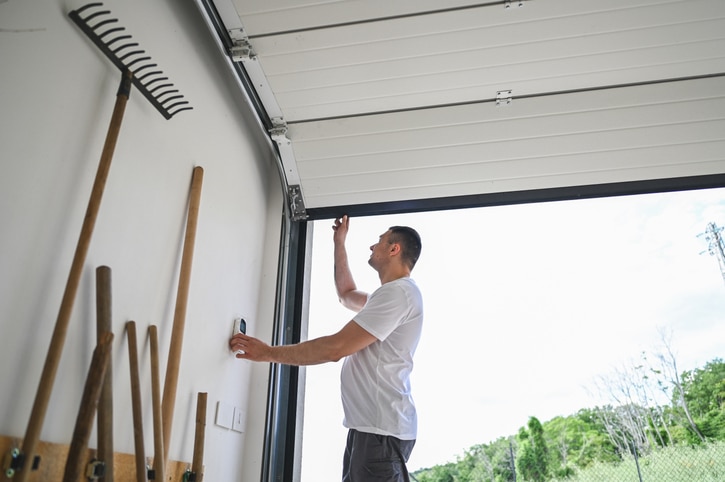 A person in a white t-shirt opens a garage door while standing next to a wall with gardening tools.