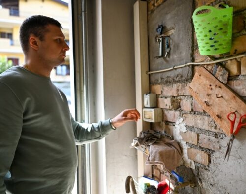 A man in a green sweatshirt stands indoors by a brick wall with tools and a thermostat, reaching toward the thermostat.