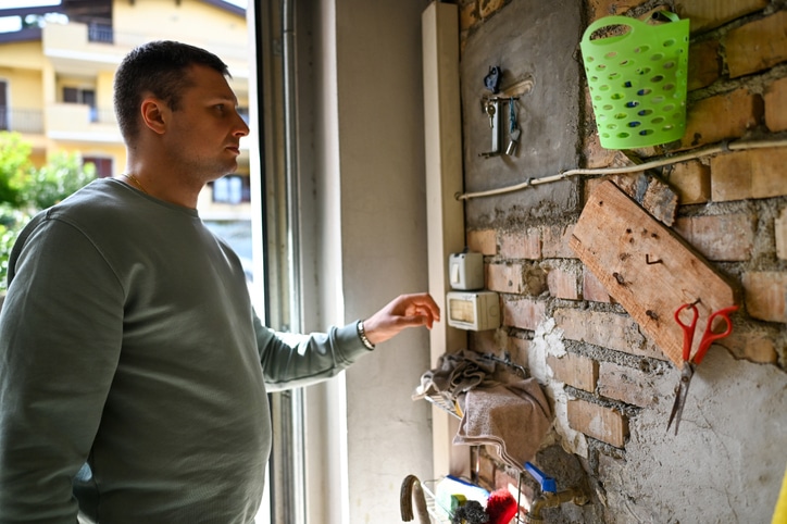 A man in a green sweatshirt stands indoors by a brick wall with tools and a thermostat, reaching toward the thermostat.