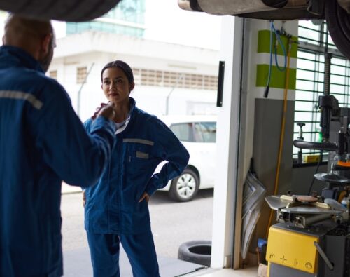 Two auto mechanics in blue uniforms are talking in a garage, with equipment and a car visible around them.