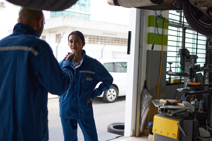 Two auto mechanics in blue uniforms are talking in a garage, with equipment and a car visible around them.