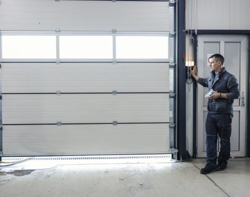 A man in work clothes stands by a control panel, operating a large white industrial garage door in a workshop or warehouse setting.