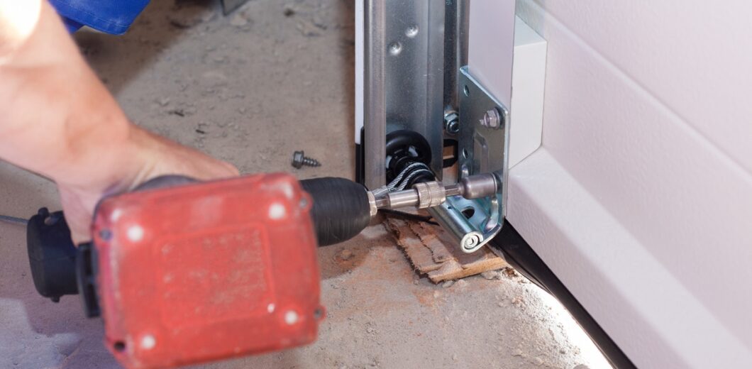 A person uses a power drill to install or repair a metal bracket at the base of a garage door track on a concrete floor.