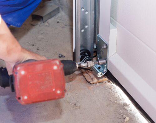 A person uses a power drill to install or repair a metal bracket at the base of a garage door track on a concrete floor.
