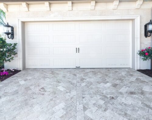 Closed white garage door with two handles, bordered by two black lantern lights, shrubs, and flowering plants on either side, with patterned stone driveway in front.