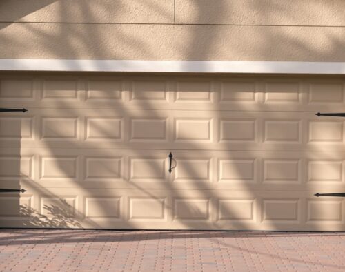 Closed beige garage door with rectangular panels, black decorative hardware, and shadows of tree branches on the surface. Brick-patterned driveway in front.
