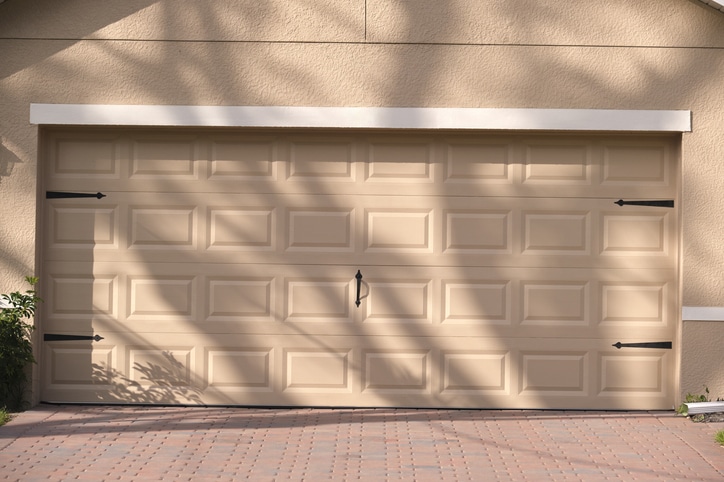 Closed beige garage door with rectangular panels, black decorative hardware, and shadows of tree branches on the surface. Brick-patterned driveway in front.