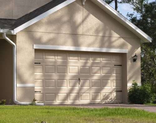 A beige single-car garage with a closed sectional door attached to a house, surrounded by greenery and a paved driveway.
