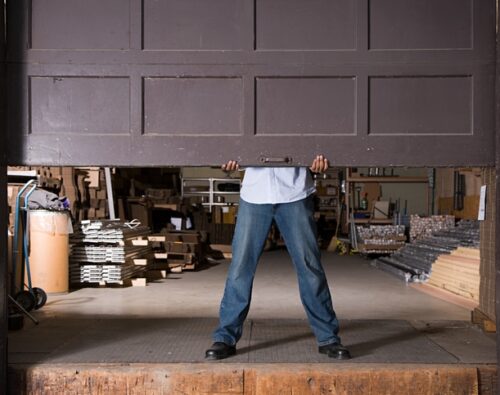 A person in jeans and a shirt lifts a large garage door in a warehouse filled with various stacked materials and equipment.