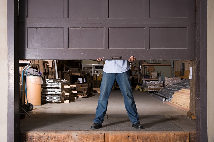A person in jeans and a shirt lifts a large garage door in a warehouse filled with various stacked materials and equipment.