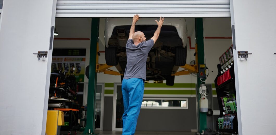 A person in blue pants opens a garage door, with a car elevated on a lift inside an automotive workshop.