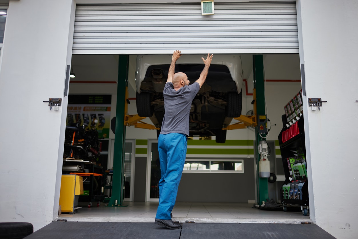 A person in blue pants opens a garage door, with a car elevated on a lift inside an automotive workshop.
