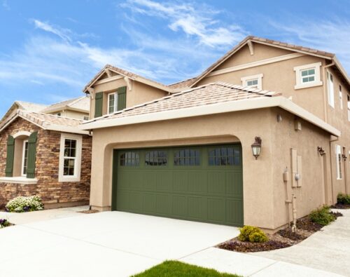 Two-story beige suburban house with a green garage door, stone accents, manicured lawn, and clear blue sky in the background.