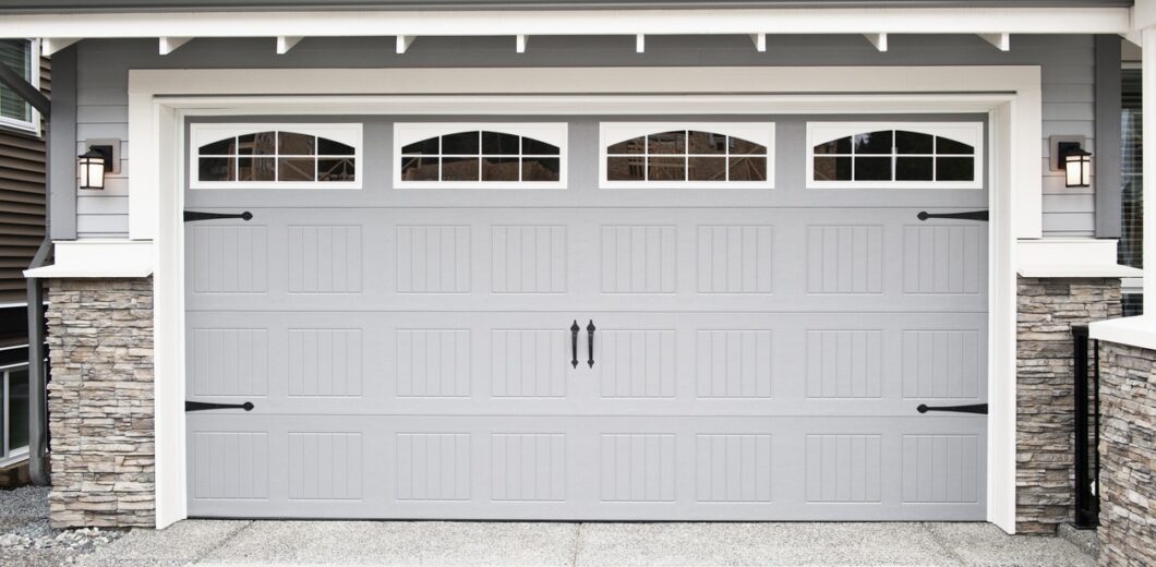 Closed gray garage door with decorative handles, window panels at the top, and stone accents on the sides of a residential house exterior.