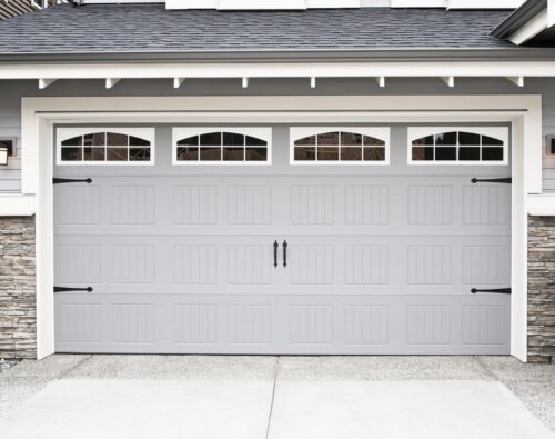 Closed gray garage door with decorative handles, window panels at the top, and stone accents on the sides of a residential house exterior.