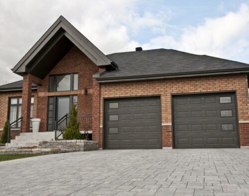A modern brick house with two dark garage doors, a paved driveway, front steps, and minimal landscaping under a partly cloudy sky.
