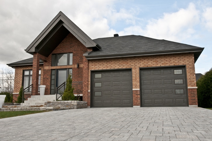 A modern brick house with two dark garage doors, a paved driveway, front steps, and minimal landscaping under a partly cloudy sky.